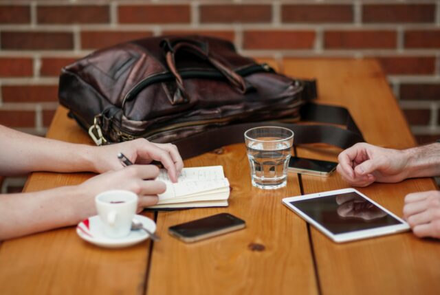 Deux personnes assises à une table avec leurs téléphones cellulaires, un iPad, un bloc-notes, un sac d’ordinateur et des boissons.